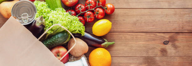 Paper grocery bag with fresh vegetables, fruits, milk and canned goods on wooden backdrop. Food delivery, shopping, donation concept. Healthy food background. Flat lay, copy space.
